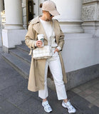 Woman in beige trench coat and white woven leather crossbody bag, wearing white sneakers and cap on city street