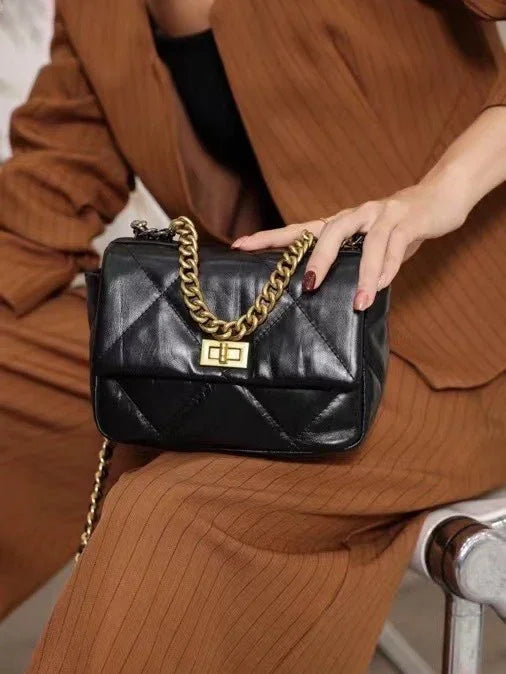 Black quilted leather shoulder bag with gold chain strap and lock, held by woman in brown suit.