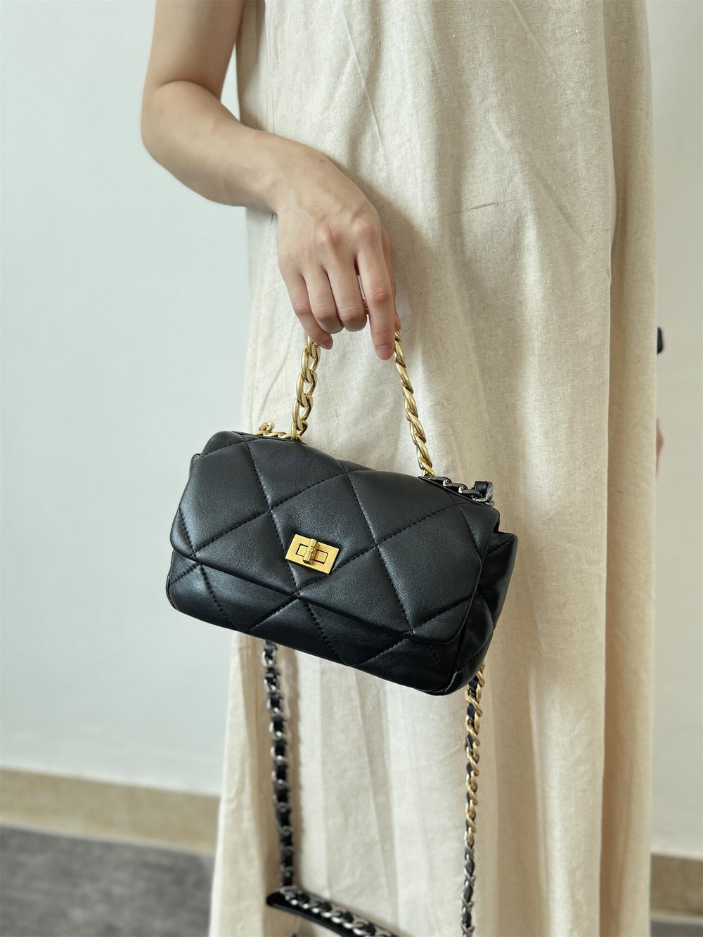 Black quilted leather shoulder bag with gold chain strap and turn-lock clasp held by a woman in a beige dress