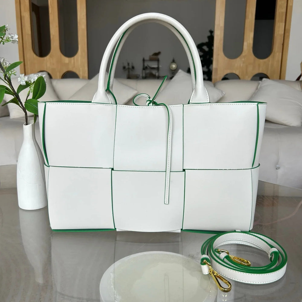 White woven leather tote bag with green trim, top handles, and detachable shoulder strap on a table.