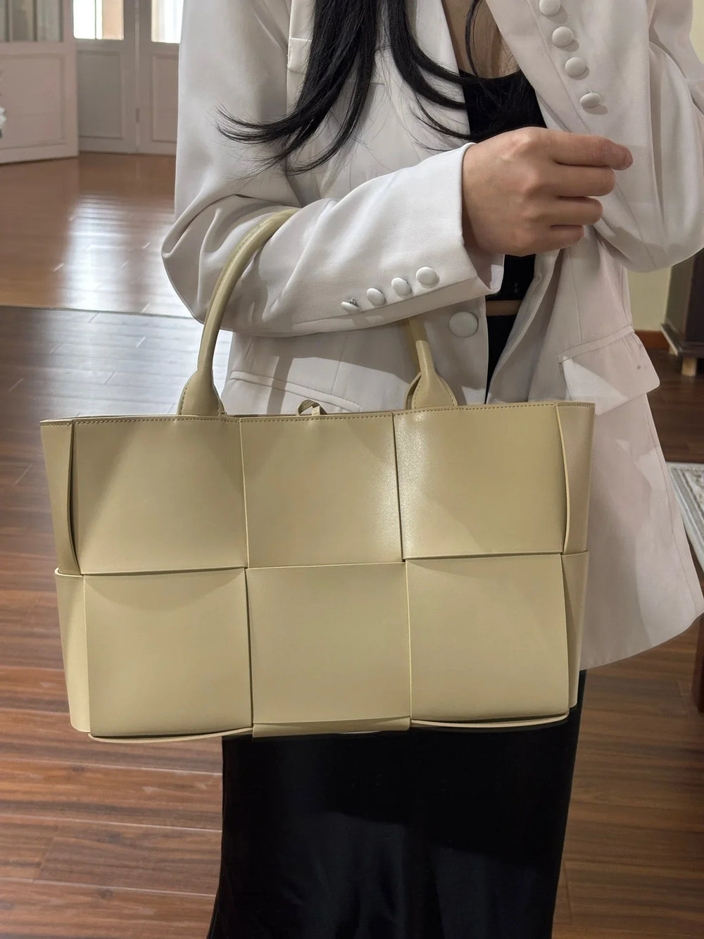 Woman in white blazer holding large beige woven leather tote bag with top handles indoors