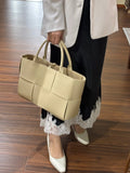 Woman holding a large beige woven leather tote bag with top handles, wearing a white blazer, lace-trimmed black skirt, and white shoes on wooden floor