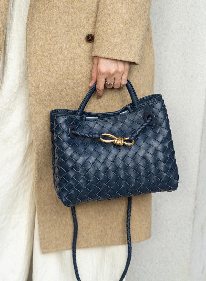 Navy blue woven lambskin leather shoulder bag with gold metal buckle, held by person in beige coat.