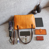 Large brown leather tote bag with dark handles on a sofa, surrounded by daily essentials like a wallet, tablet, phone, umbrella, hairbrush, perfume, and sanitizer.