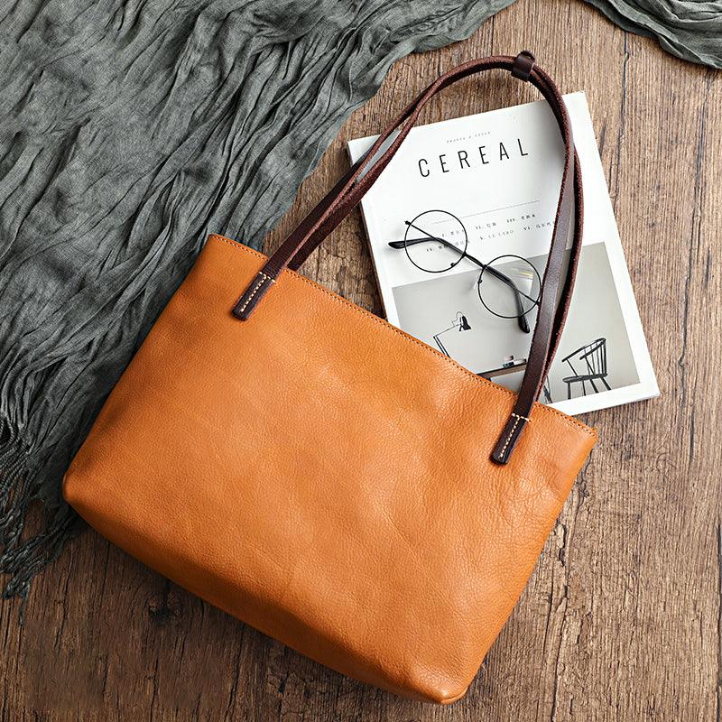 Large tan leather tote bag with dark handles on wooden surface, magazine and glasses nearby
