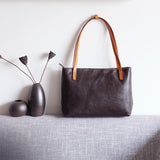 Black full grain leather tote bag with brown straps on a grey sofa, minimalist decor