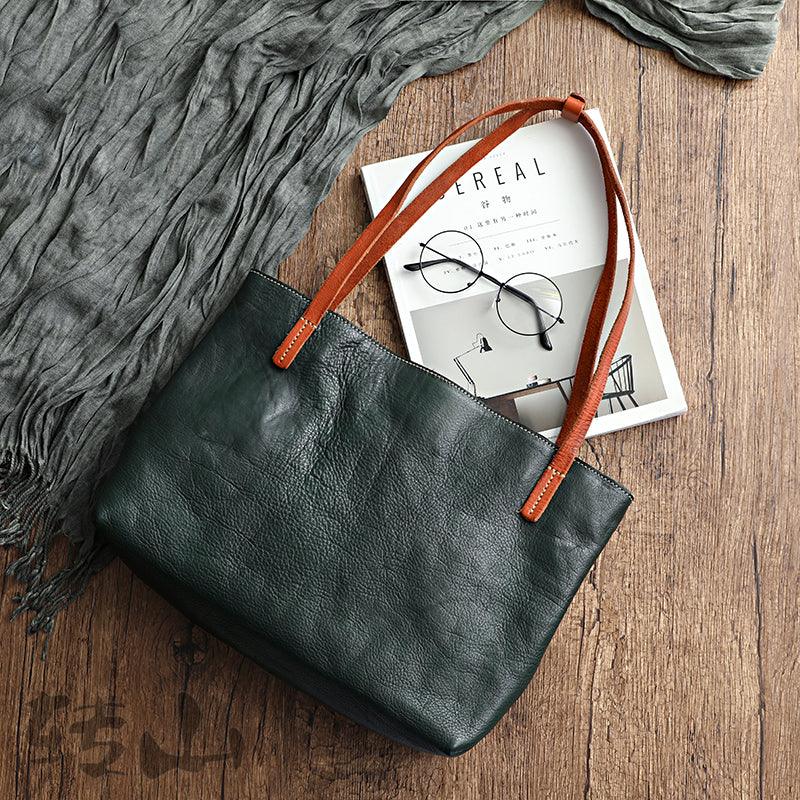 Black full grain leather tote bag with brown handles on wooden surface, beside magazine and glasses