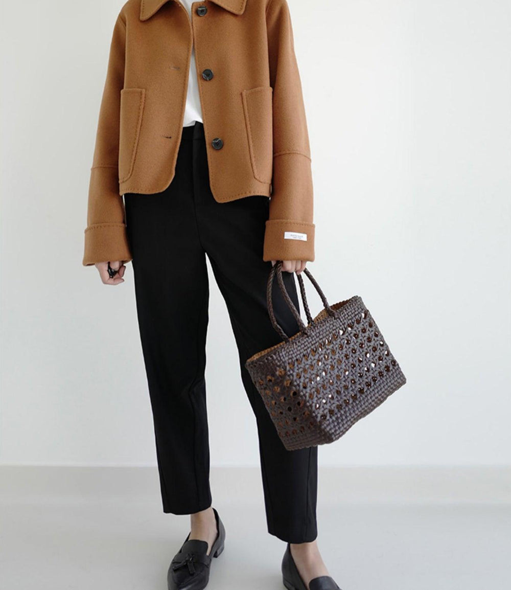 Woman holding a brown woven leather tote bag, styled with a tan jacket and black pants