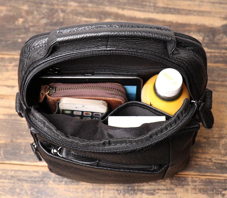 Open black leather men's crossbody shoulder bag with phone, wallet, drink, and book inside on wooden table