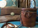 Handcrafted brown leather clutch with decorative metal accent and round leather shoulder bag on patterned fabric background.