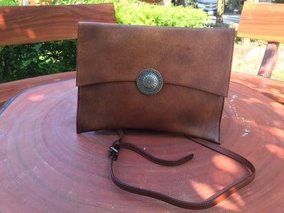 Handcrafted brown leather clutch bag with shoulder strap and decorative metal button, displayed outdoors.