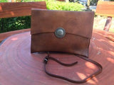 Handcrafted brown leather clutch bag with shoulder strap and decorative metal button, displayed outdoors.