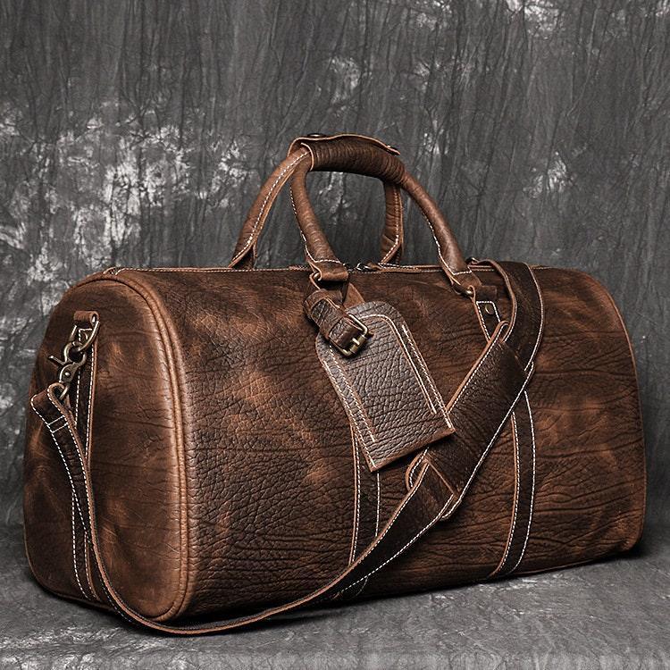 Full grain brown leather duffle bag with detachable strap and ID tag on textured background