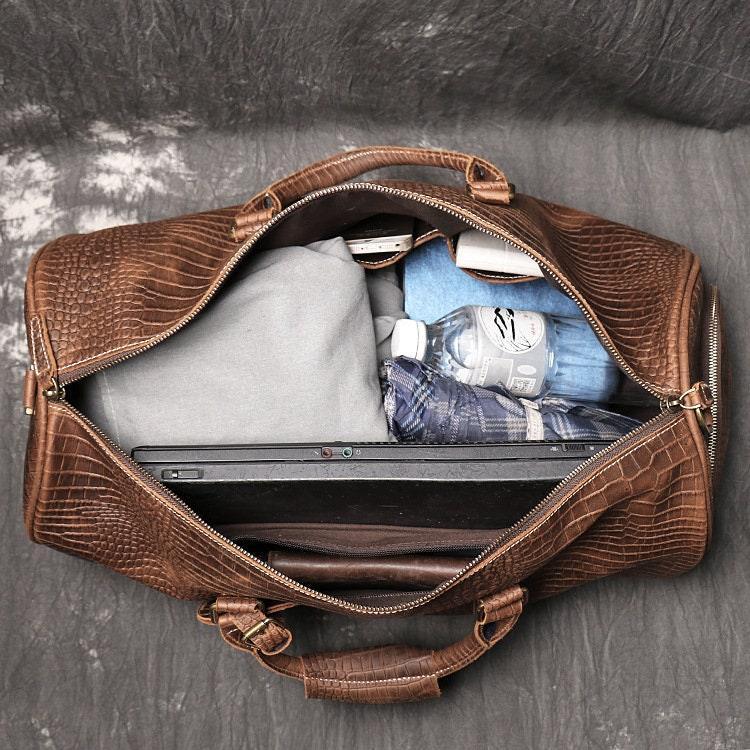 Open brown full-grain leather duffle bag filled with clothes, laptop, and a water bottle
