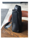 Black full grain leather shoulder bag with wide strap on a wooden table, crossbody style