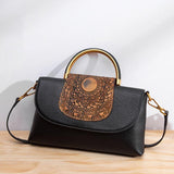 Black leather handbag with embossed brown mandala flap and gold metal handle on wood surface