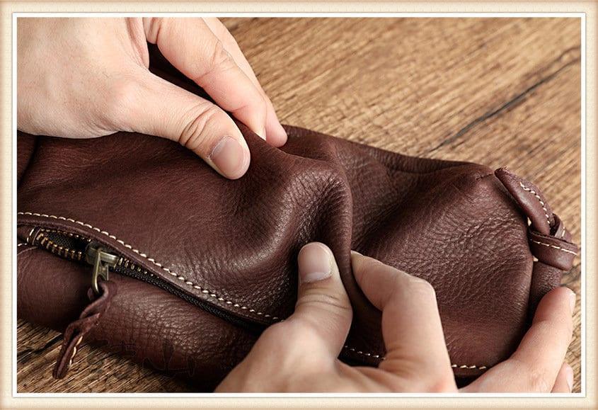 Hands squeezing a small brown cowhide leather shoulder bag on a wooden surface