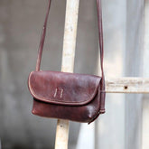 Brown cowhide leather shoulder bag with flap, handmade, vintage style, hanging on a wooden ladder.
