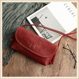 Red cowhide leather shoulder bag with strap on wood surface, beside magazine and eyeglasses