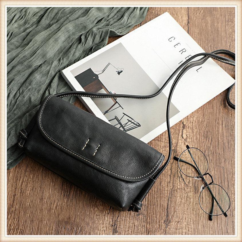 Black cowhide leather shoulder bag with white stitching on wooden surface beside glasses and magazine