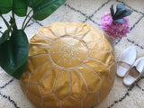 Light yellow Moroccan leather pouf with geometric stitching on a patterned rug, next to slippers and pink flowers