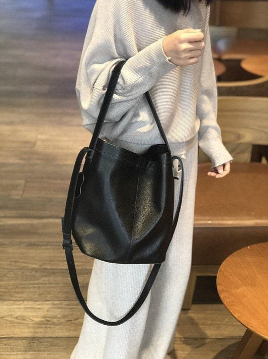 Large black leather bucket bag with adjustable shoulder strap, worn by woman indoors