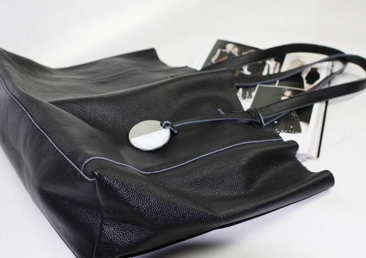 Black full grain leather tote bag with long handles and round metal charm on white background