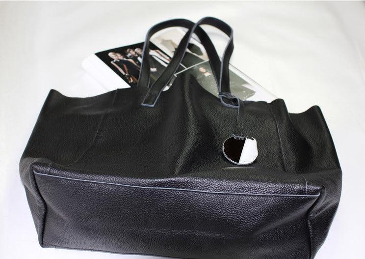 Black full grain leather tote bag with double handles and attached tag on white background