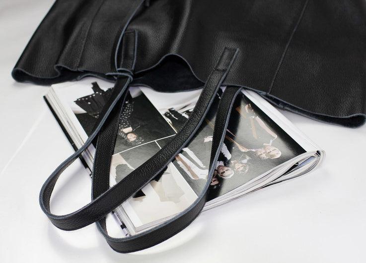 Black full grain leather tote bag with long straps, open on top of a fashion magazine