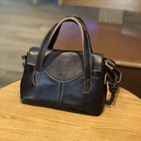 Small black minimalist leather handbag with top handles and crossbody strap on a wooden table