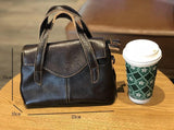 Small black leather crossbody handbag with top handles, shown on a table beside a coffee cup, size 23x15x10cm.