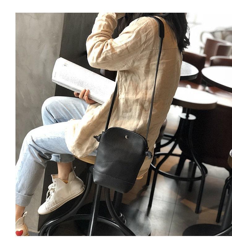 Woman seated in a cafe wearing a tan shirt and jeans, carrying a black leather crossbody bag