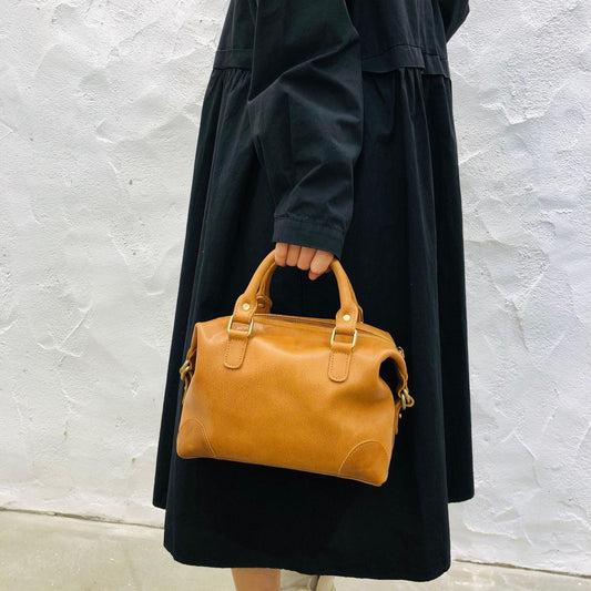 Woman holding a vintage tan leather Boston bag with top handles against a white wall background