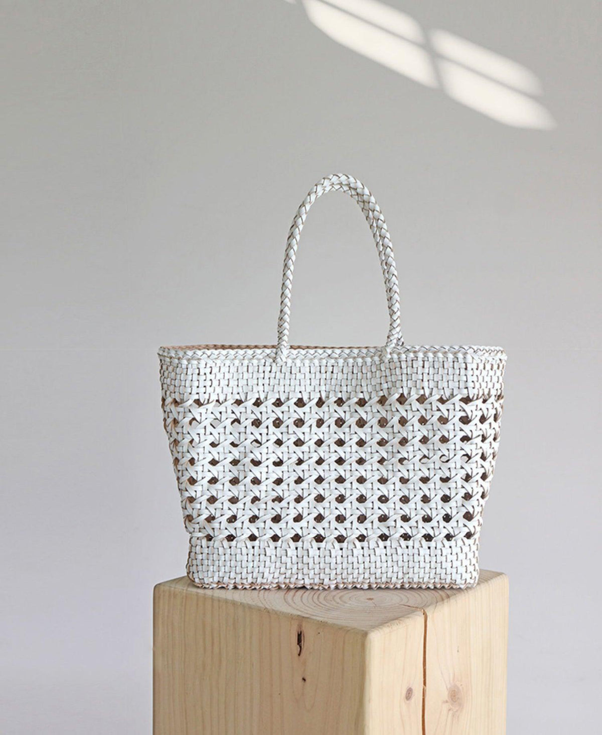 White hand-woven genuine leather tote bag with cuboid shape and braided handles on wood block