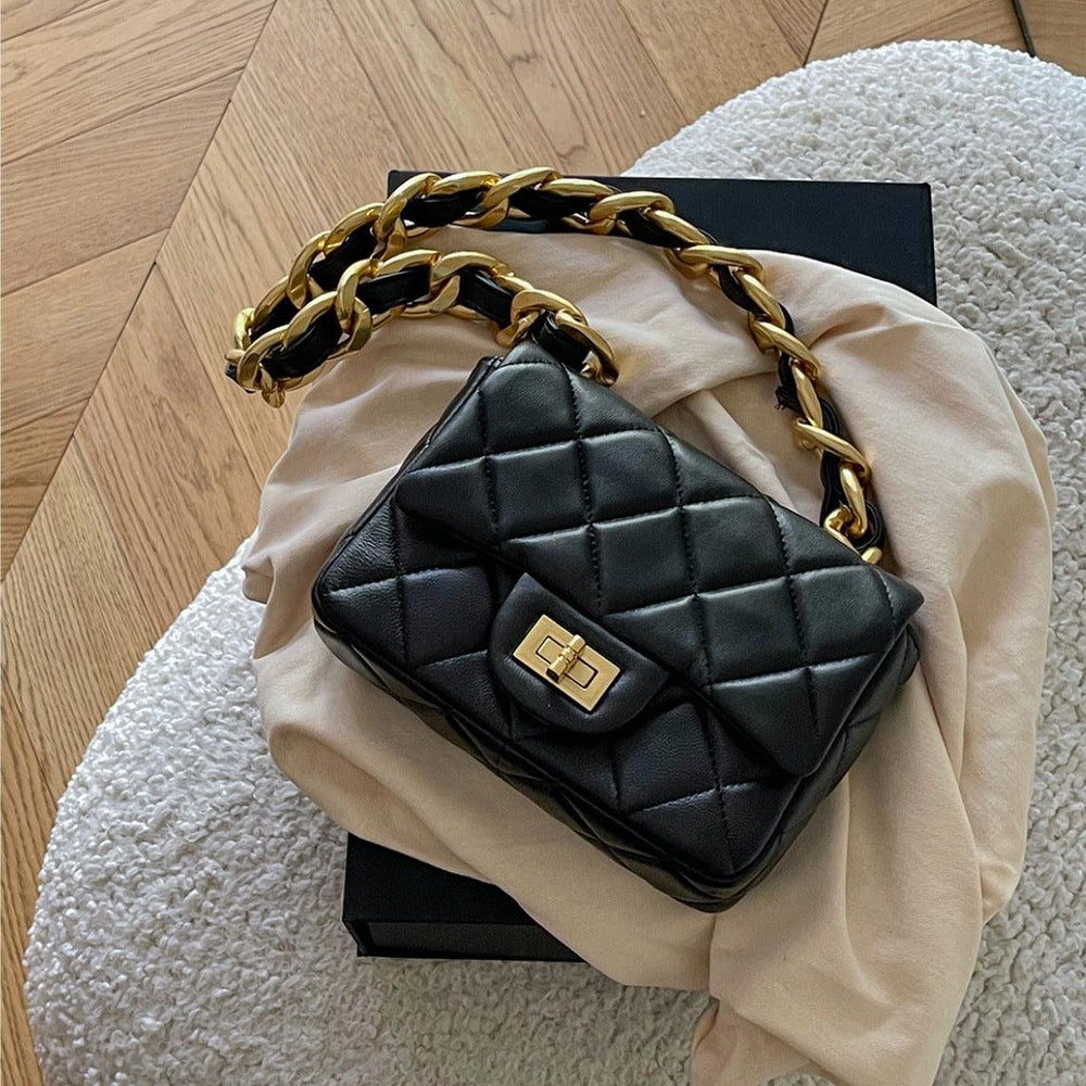 Black quilted lambskin leather shoulder bag with gold chain strap and turn-lock clasp on a beige fabric and black box.