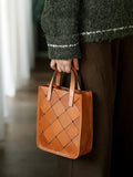 Handcrafted brown cowhide leather woven handbag with top handles, held by a person indoors