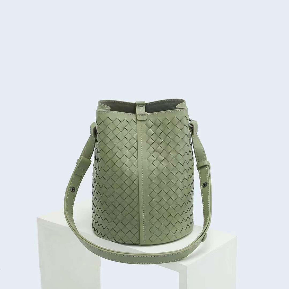 Sage green woven lambskin leather bucket bag with adjustable shoulder strap on white pedestal