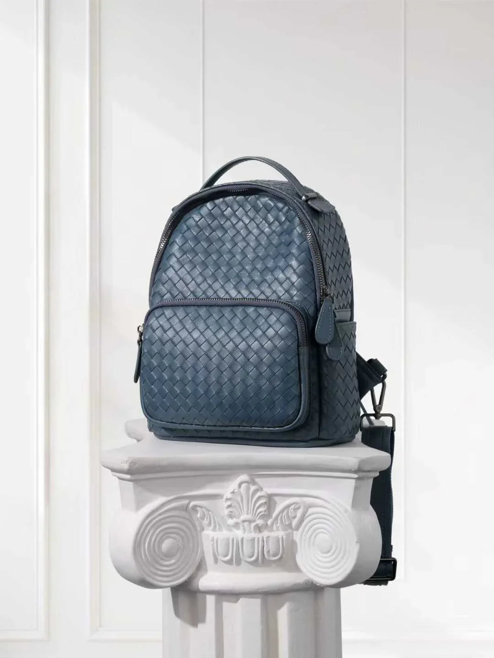 Woven lambskin leather small backpack in blue with top handle and front pocket on a white pedestal