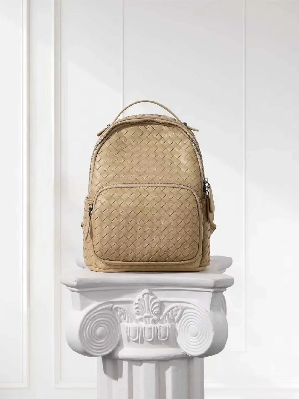 Beige handwoven lambskin leather backpack with top handle, displayed on a white pedestal.