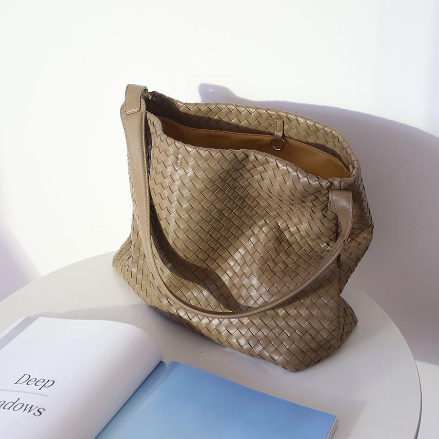 Beige woven leather tote bag with shoulder strap on a white table beside an open book