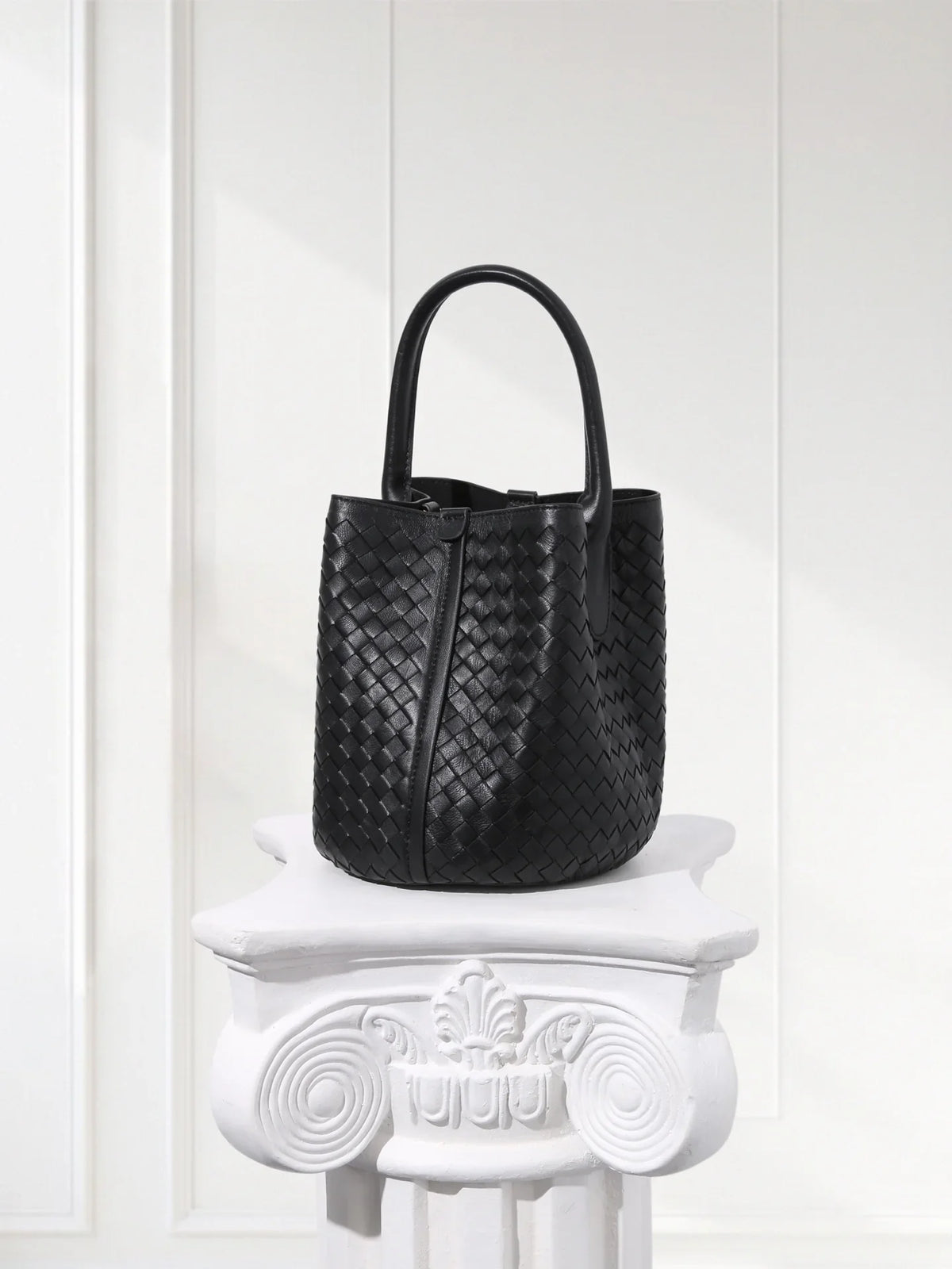 Black handwoven lambskin leather bucket bag with top handles on a white pedestal