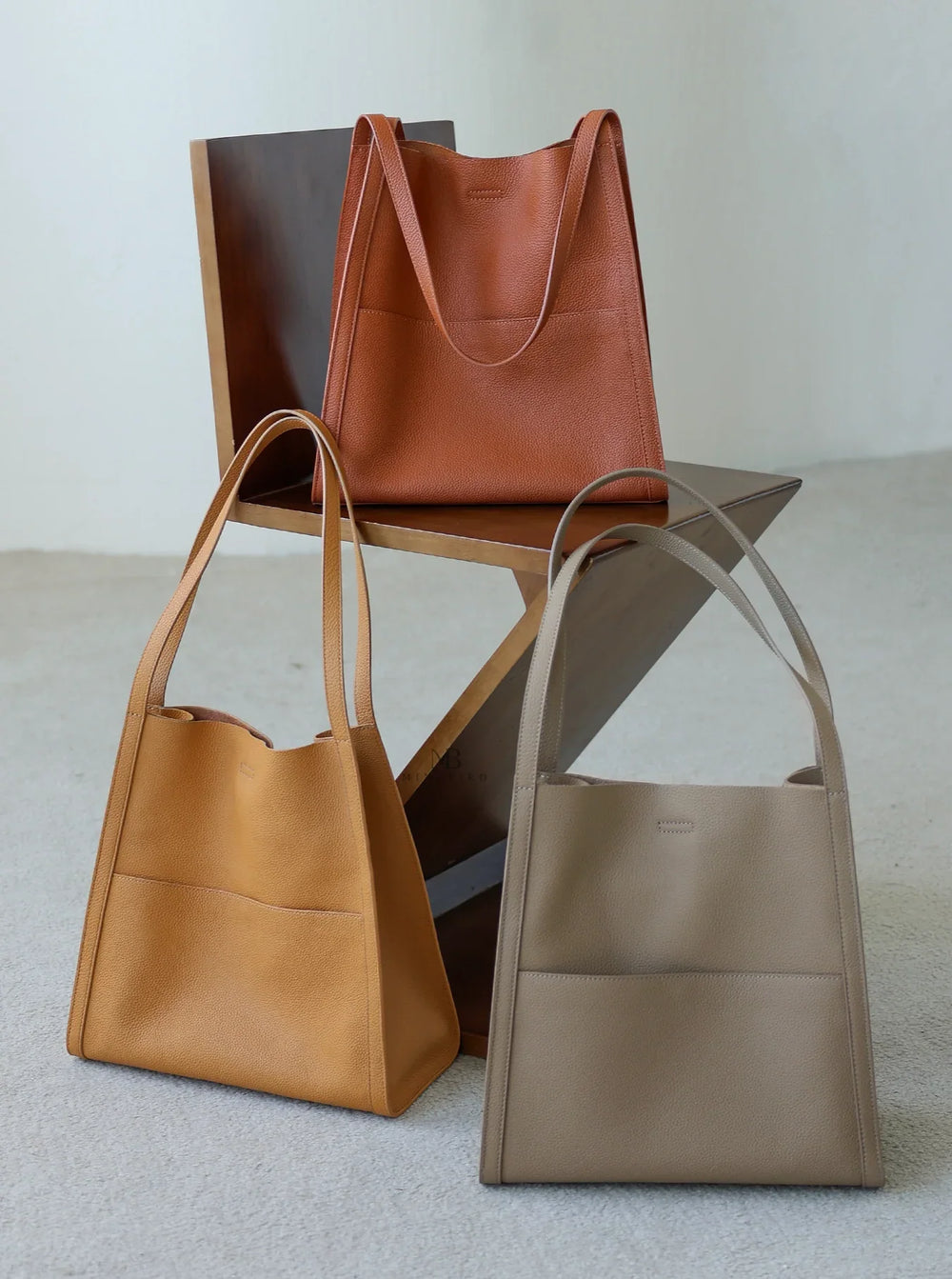 Three minimalist cowhide leather tote bags in tan, camel, and beige on a modern chair.