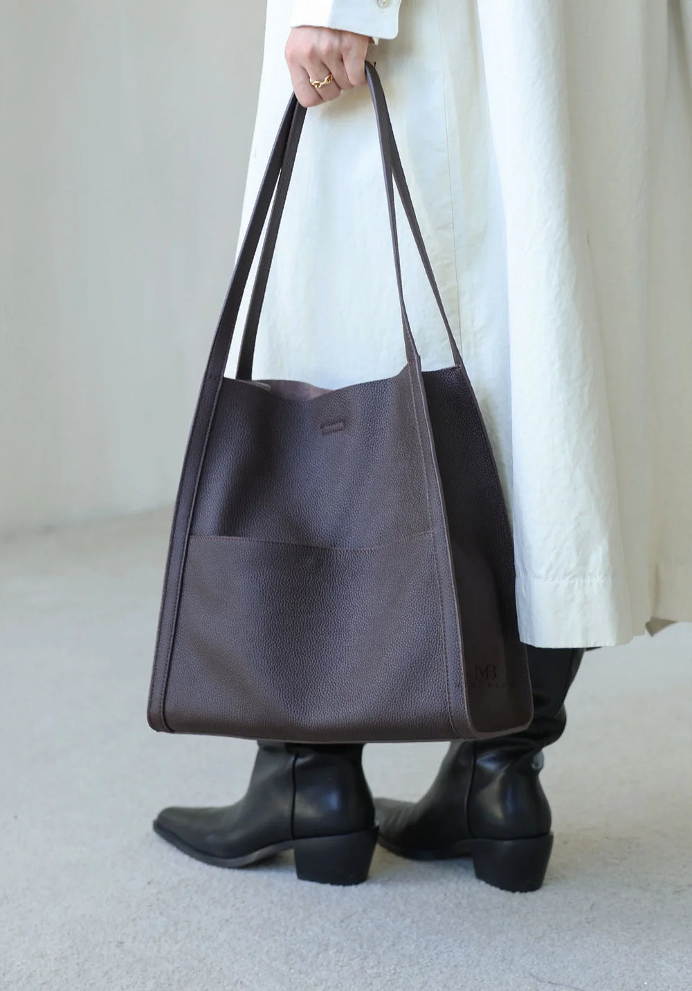 Woman holding large brown grain cowhide leather tote bag with long straps, wearing boots and a light coat.
