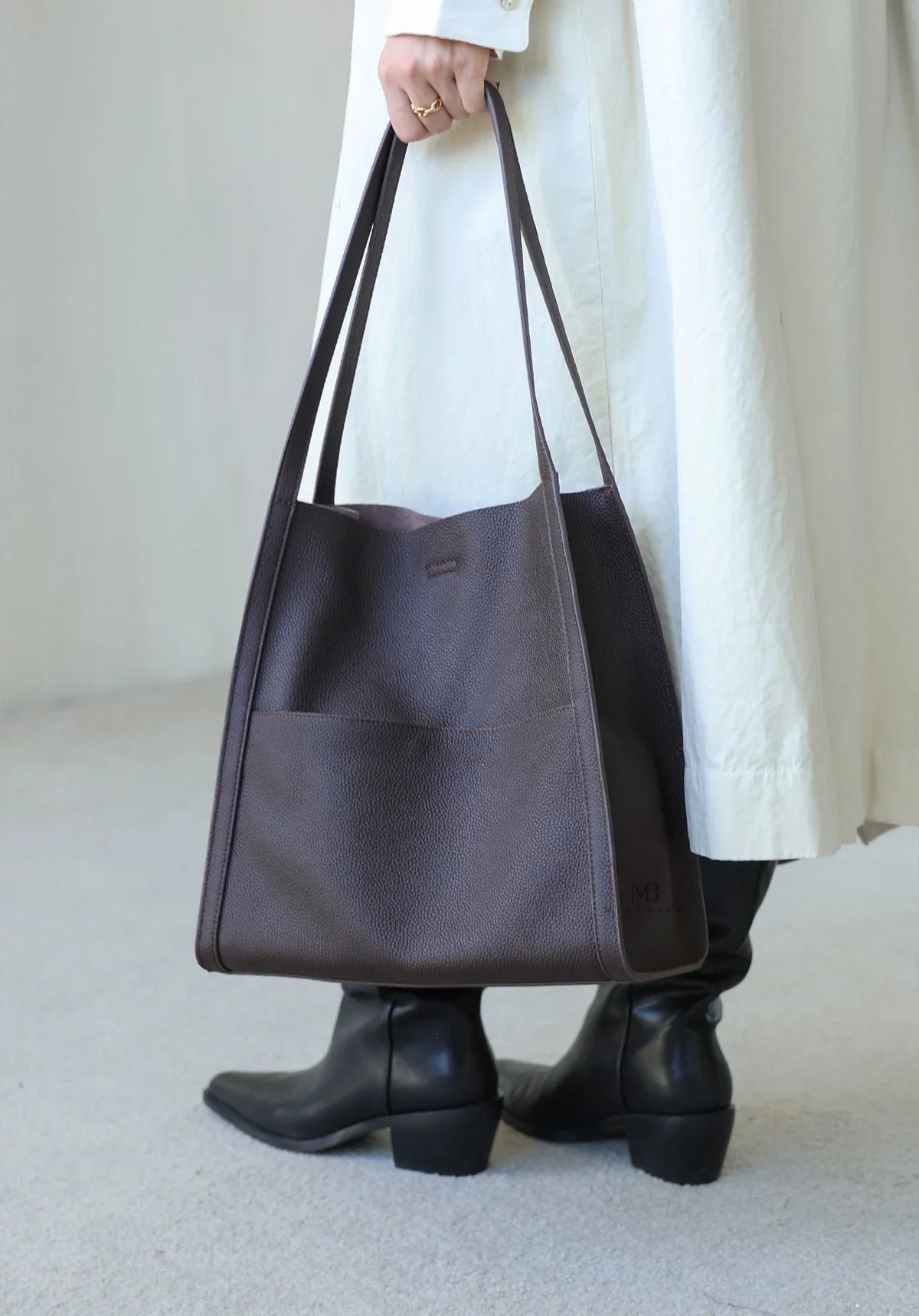 Woman holding large brown grain cowhide leather tote bag with long straps, wearing boots and a light coat.
