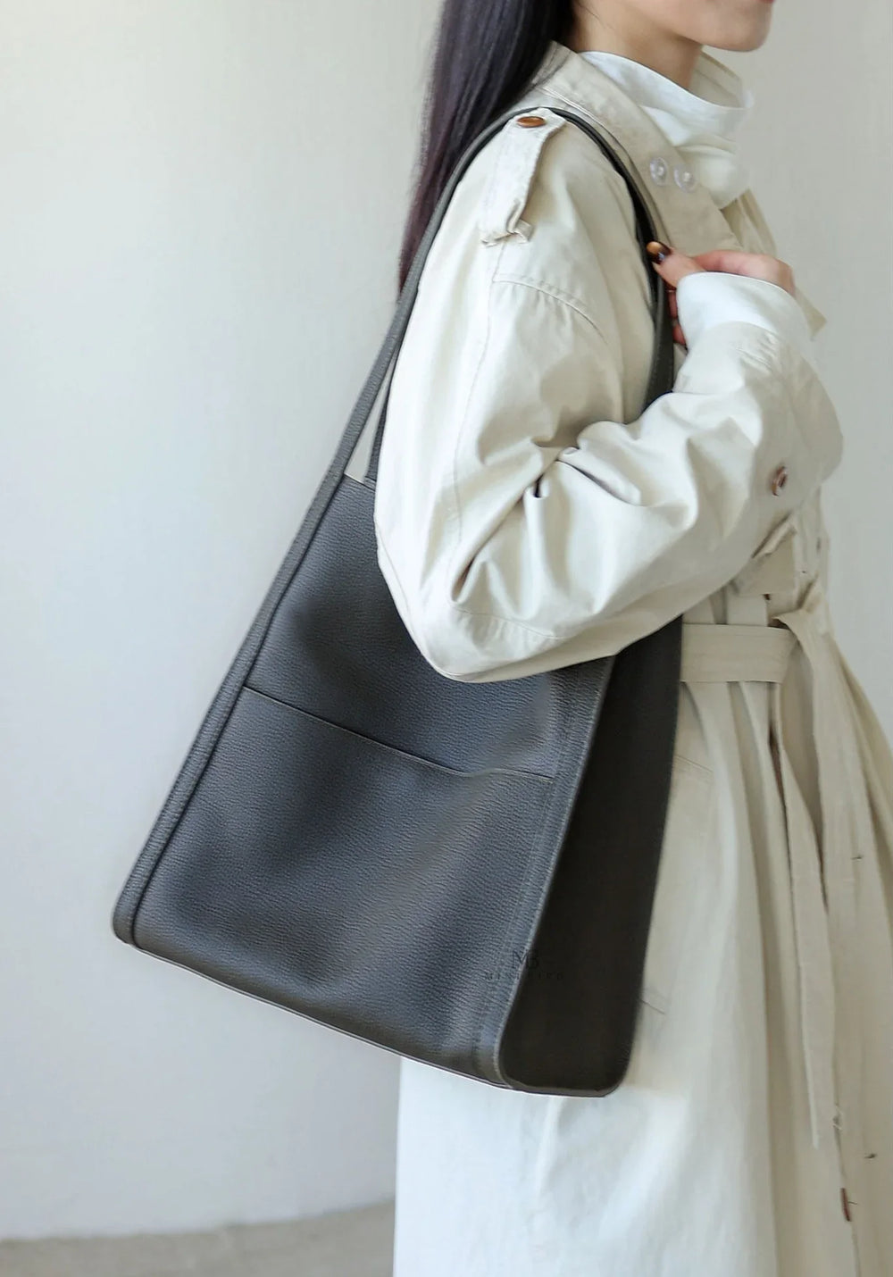Minimalist dark grain cowhide leather tote bag with shoulder strap, worn by woman in light trench coat