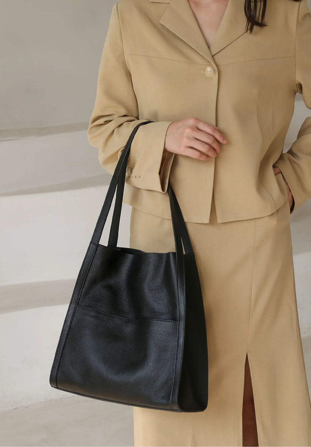 Minimalist black grain cowhide leather tote bag with double shoulder straps, held by woman in beige suit