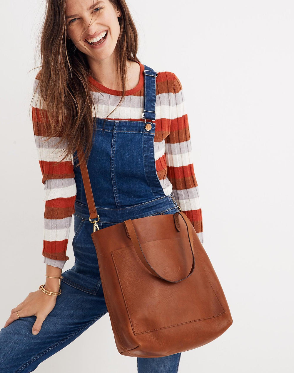Woman wearing denim overalls with a large tan full grain leather tote shoulder bag.