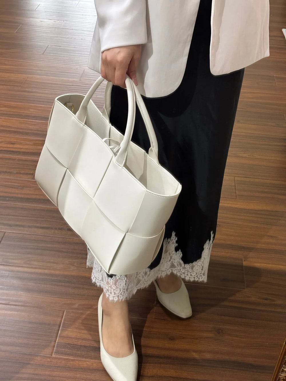Woman holding a large white woven leather tote bag with top handles, standing on wood floor