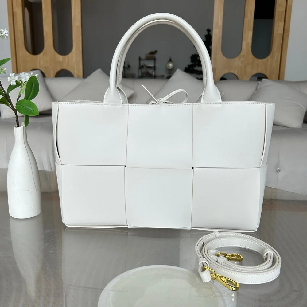 Large white woven leather tote bag with top handles and detachable strap on a glass table.