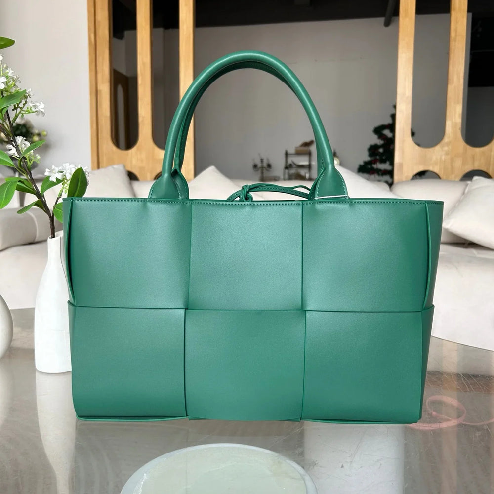 Large green woven leather tote bag with top handles on a glass table in a modern living room.